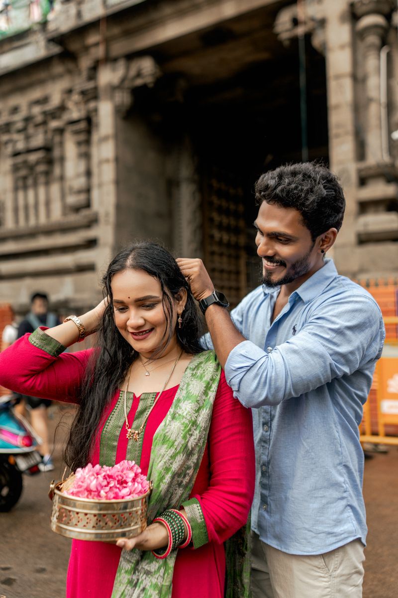 A couple shares a playful moment with flower petals in traditional Indian attire against a stone building backdrop.