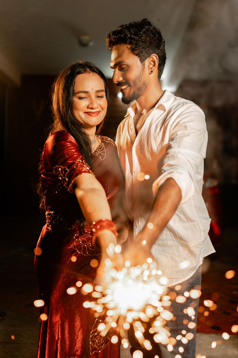 A couple in red and white outfits share an intimate moment with sparklers creating magical light effects in the dark.