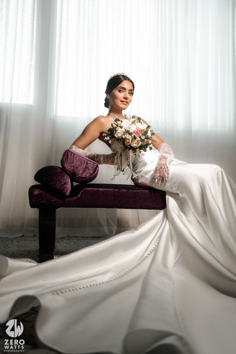 Bride in strapless white gown with lace gloves sits elegantly holding a bouquet on velvet bench.