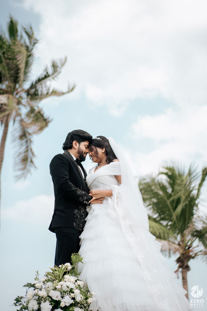 Bride and groom share romantic moment outdoors with palm trees and blue sky at beach wedding ceremony.