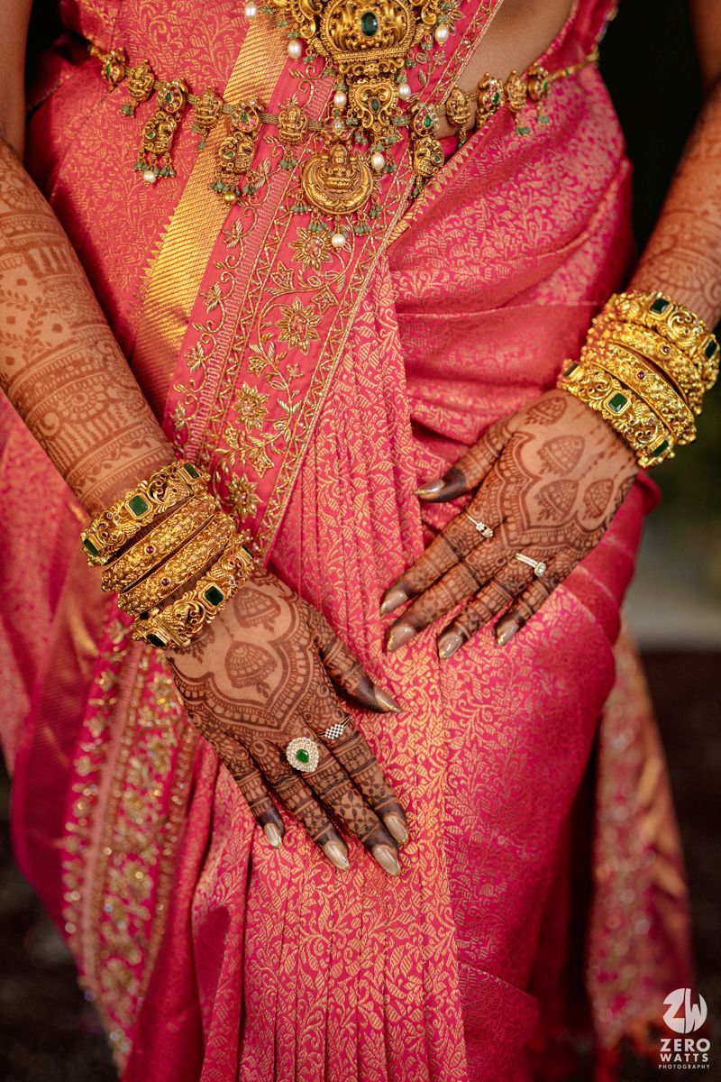 Close-up of Indian bride's hands adorned with intricate mehndi henna designs, gold bangles, rings, and a pink silk saree.