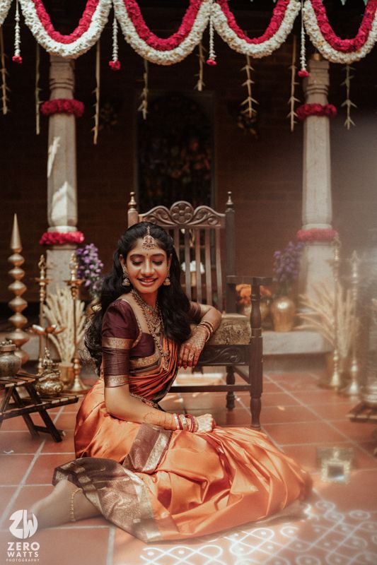 South Indian bride in orange and maroon silk saree seated gracefully amid floral decorations and traditional lamps.