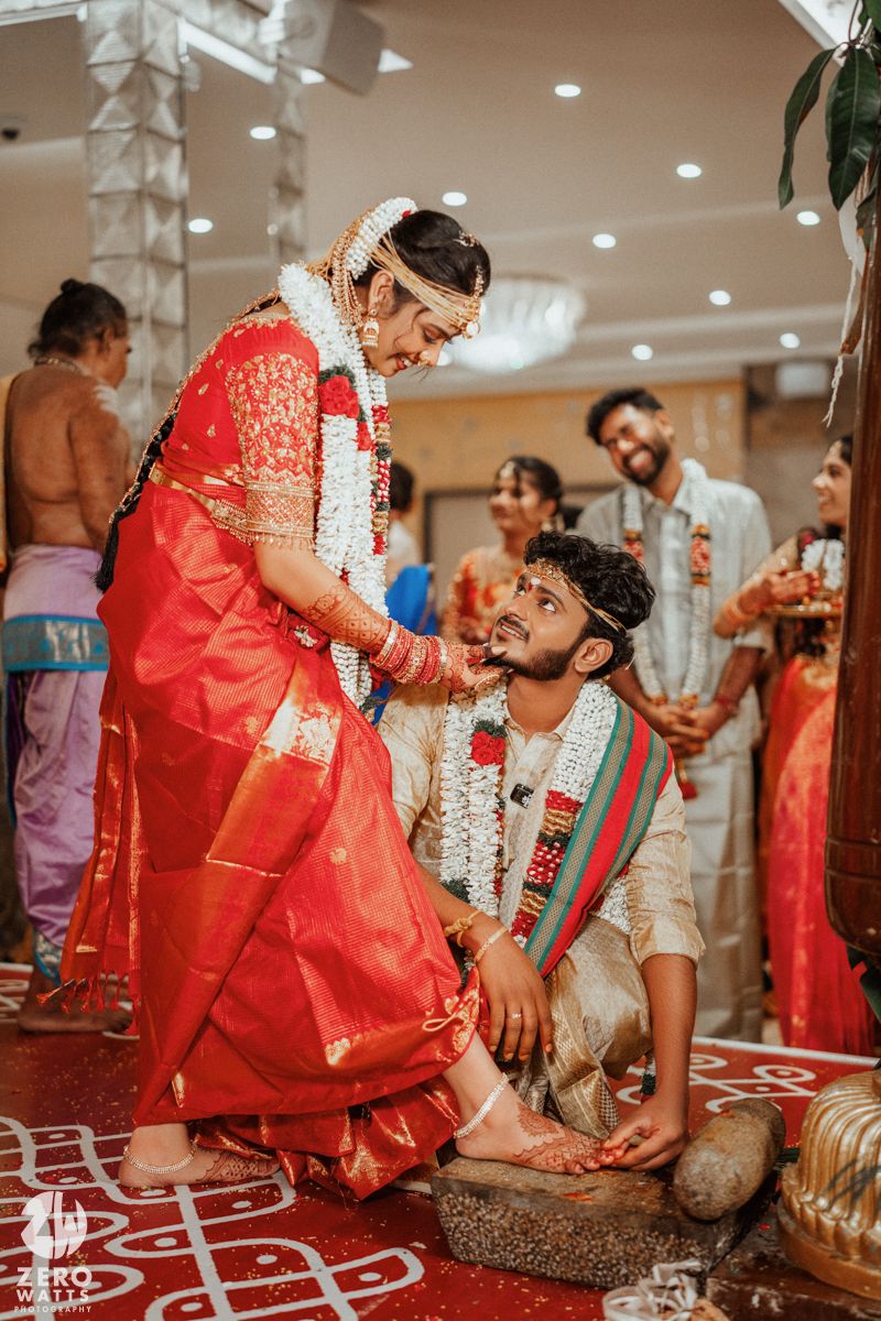 Indian wedding ceremony with bride in red saree and groom kneeling during traditional ritual.