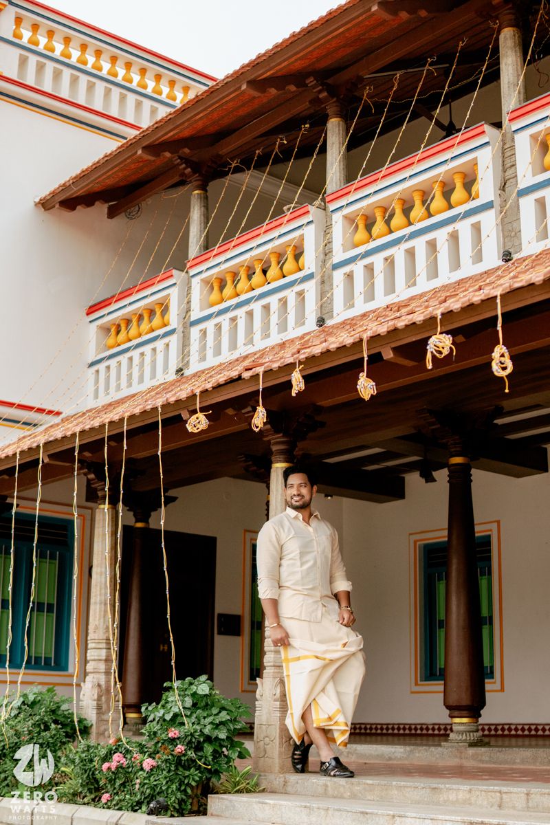 Man in traditional South Indian attire standing on steps of ornate heritage building with decorative balcony.