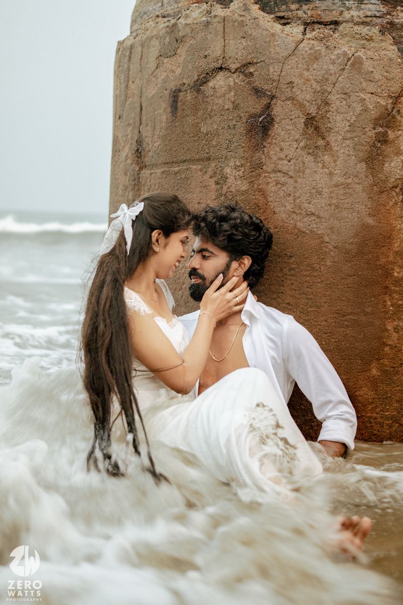 Romantic couple in white outfits sitting in ocean waves beside a rock during a beach pre-wedding photoshoot.