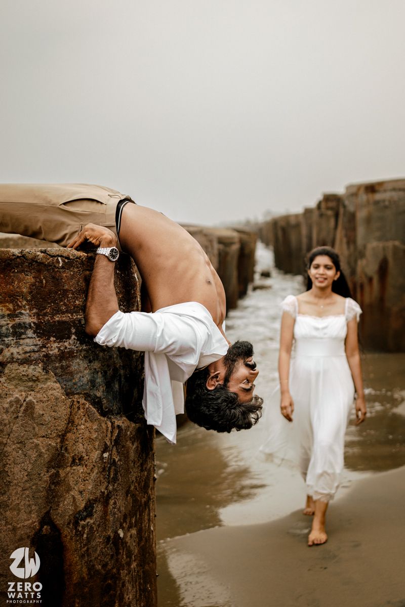Man hangs upside down from beach pier as woman in white dress walks toward him during pre-wedding shoot.