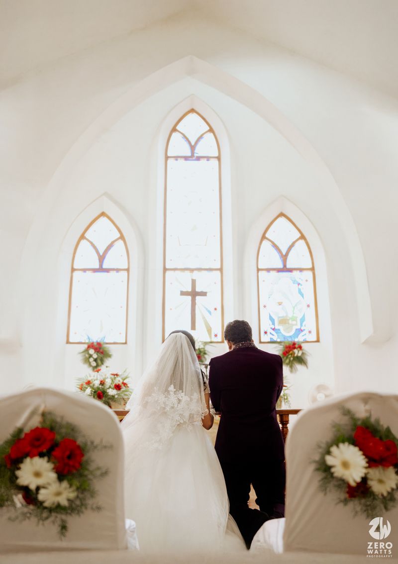 Bride and groom standing at church altar with stained glass windows and cross, viewed from behind.