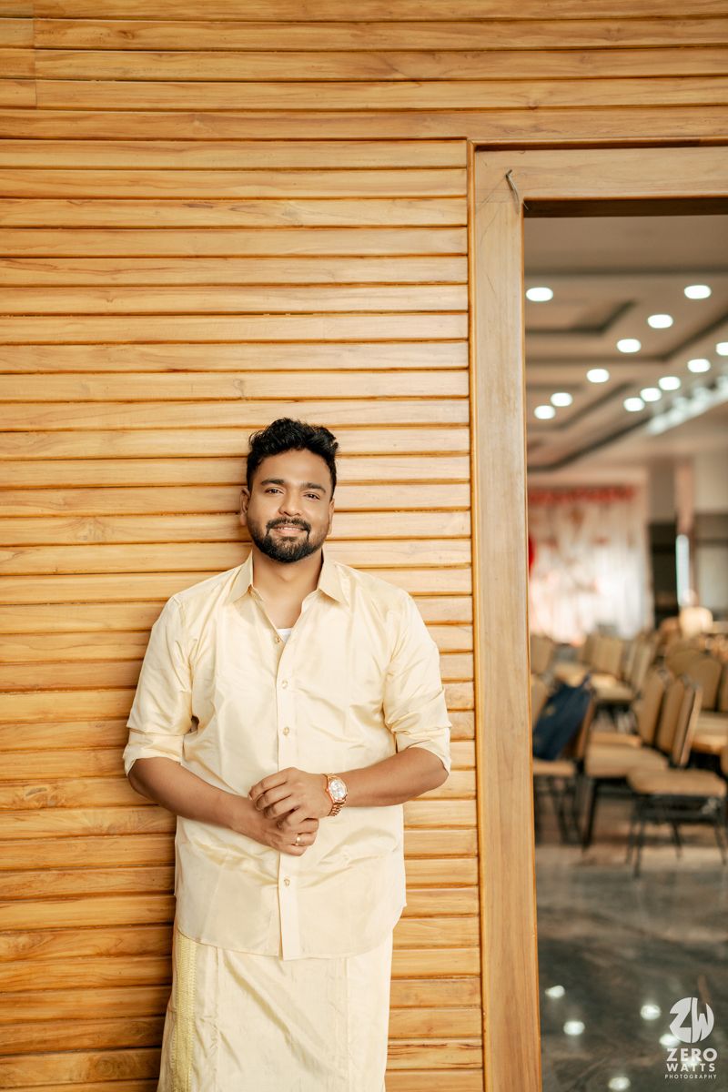 Smiling man in cream kurta leaning against wooden wall in retail store.