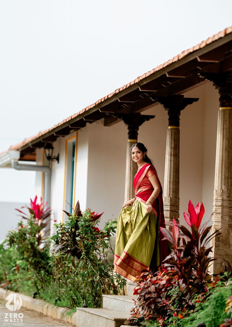 Bride in elegant red and green half saree standing on steps of a traditional South Indian heritage home.