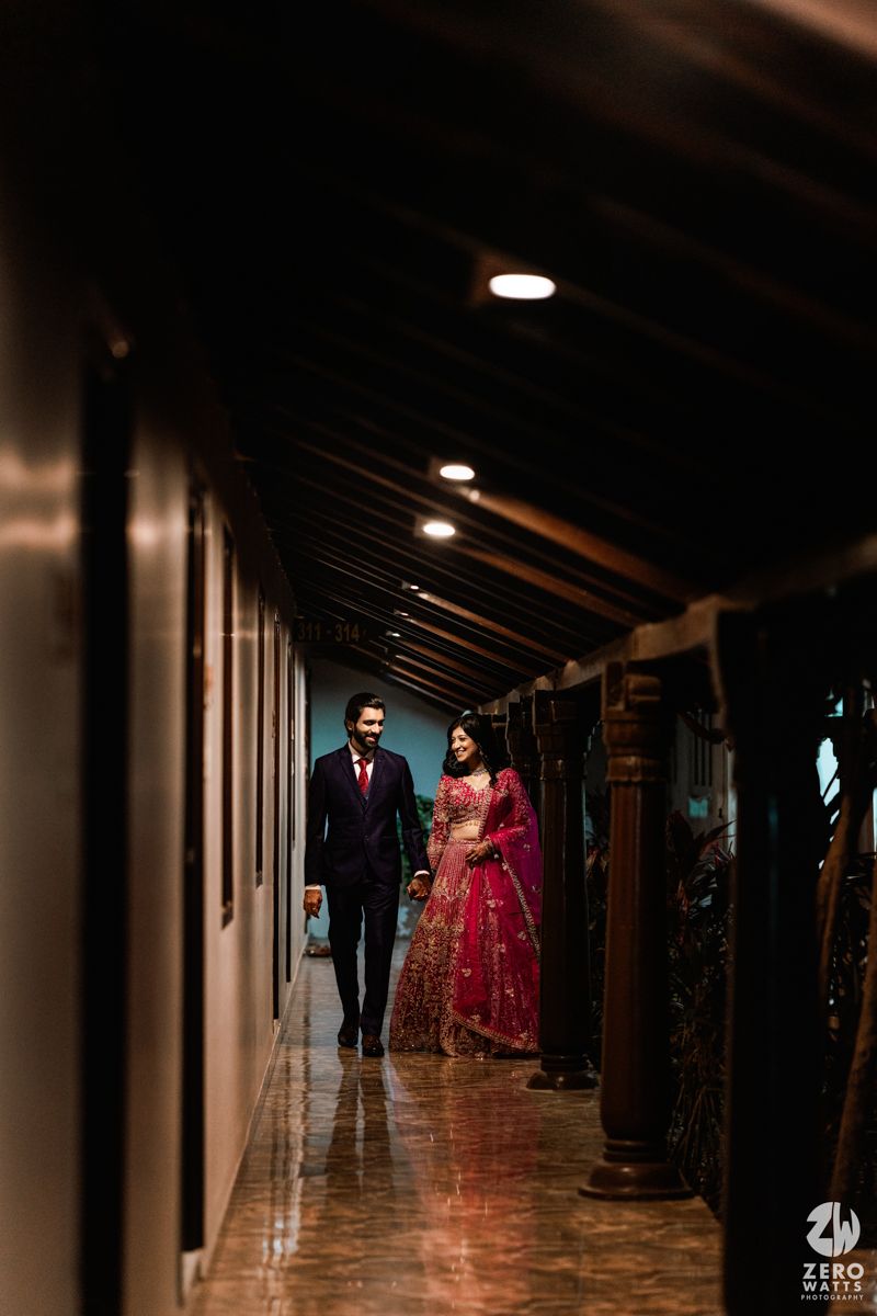 Elegant Indian couple walking hand-in-hand through a warmly lit corridor, bride in pink lehenga and groom in navy suit.