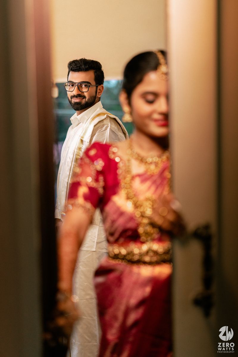 Groom in white dhoti gazing at bride in red Kanjivaram saree through doorway before wedding.