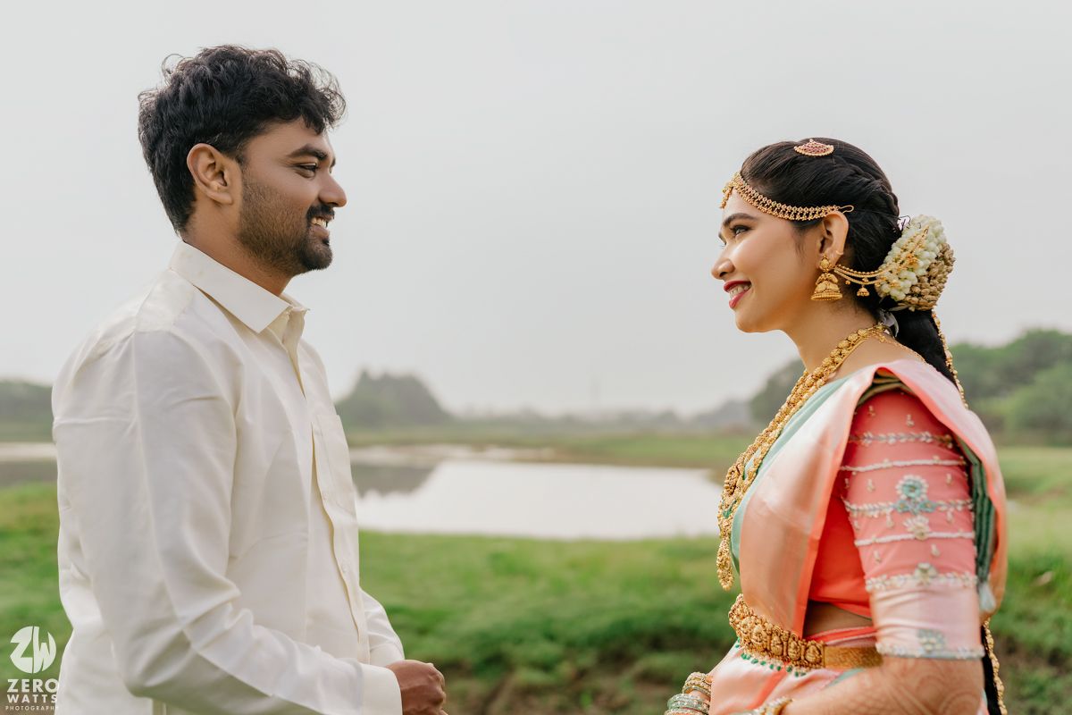 An Indian couple in traditional wedding attire share a joyful moment outdoors near a serene lake.