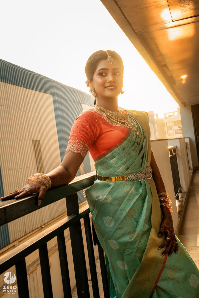 Bride in teal silk saree with orange blouse and gold jewelry poses on balcony in warm sunset light.