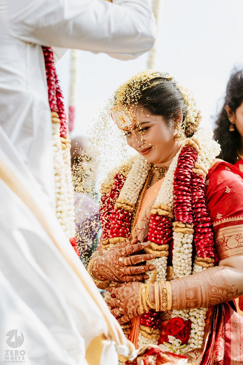Bride in red saree adorned with floral garlands during traditional South Indian wedding ceremony with rice shower.