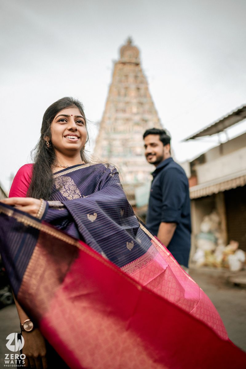 A person holds up and displays a traditional silk saree with navy blue and red colors against a temple backdrop.