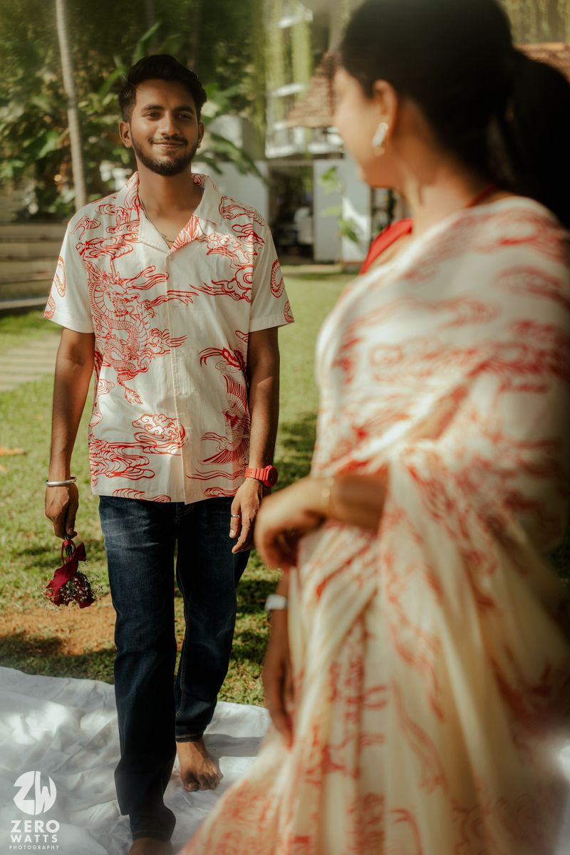 A couple in casual attire stands together outdoors, with one wearing a white and red patterned Hawaiian shirt.