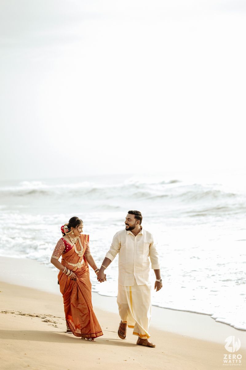 A couple in traditional Indian attire walks along a sandy beach with ocean waves in the background.