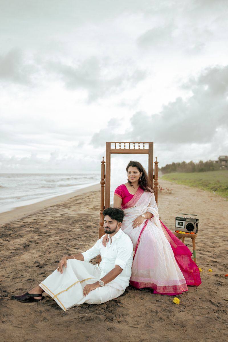 A romantic beach photoshoot featuring a couple in traditional Indian attire lounging on wooden chairs with a vintage radio.