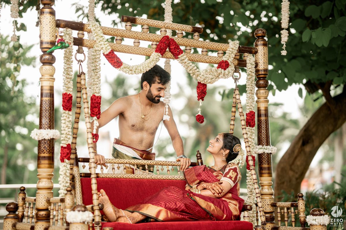 Couple posing on an ornate wooden swing decorated with red flowers and traditional Indian wedding decor.