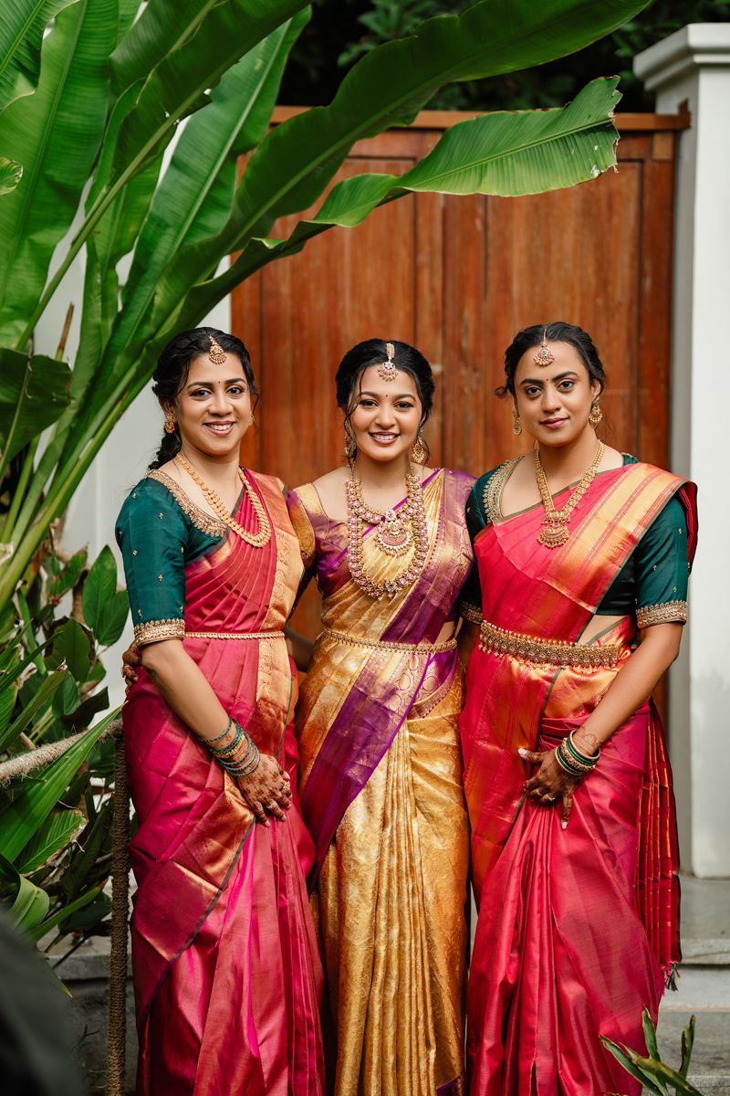 Three people in vibrant silk sarees pose together in front of tropical plants and a wooden fence.