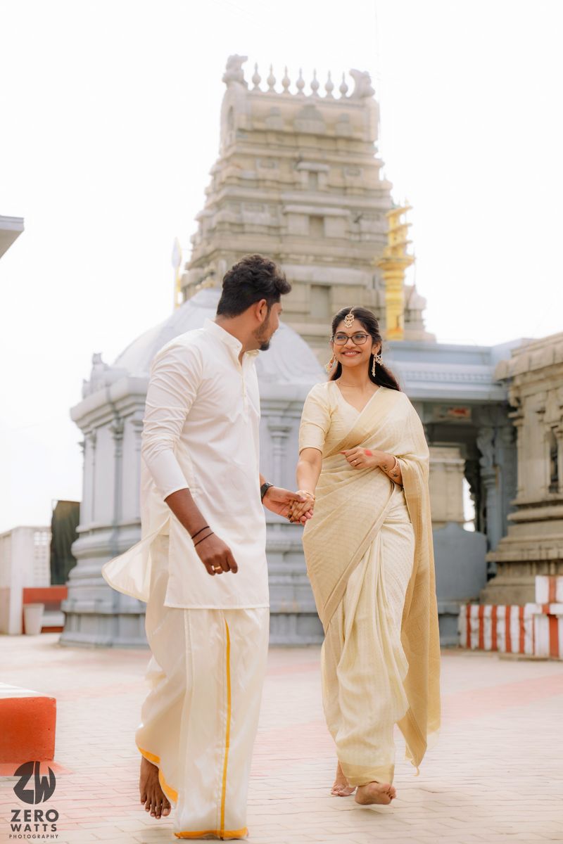 Two people in traditional white Indian attire share a moment in front of a temple with ornate architecture.