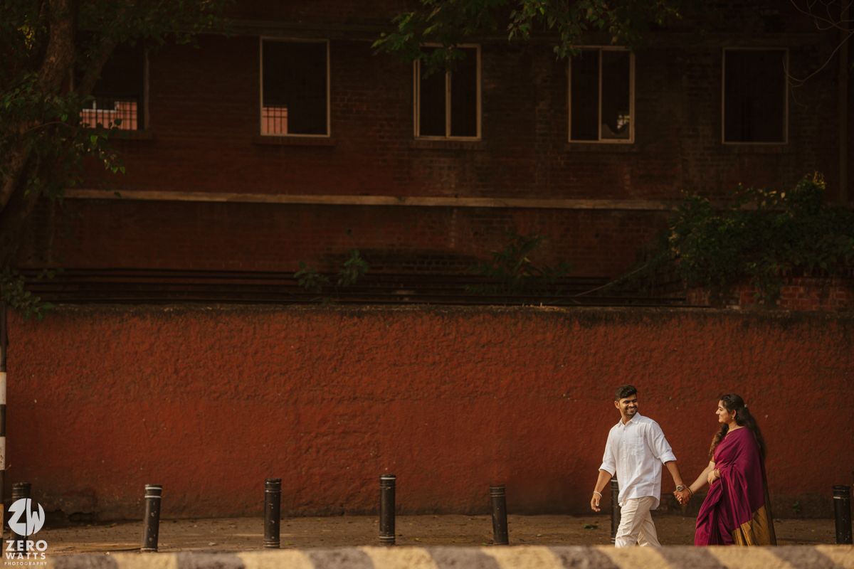 Couple walking together on a path beside a rustic brick building in warm sunlight.