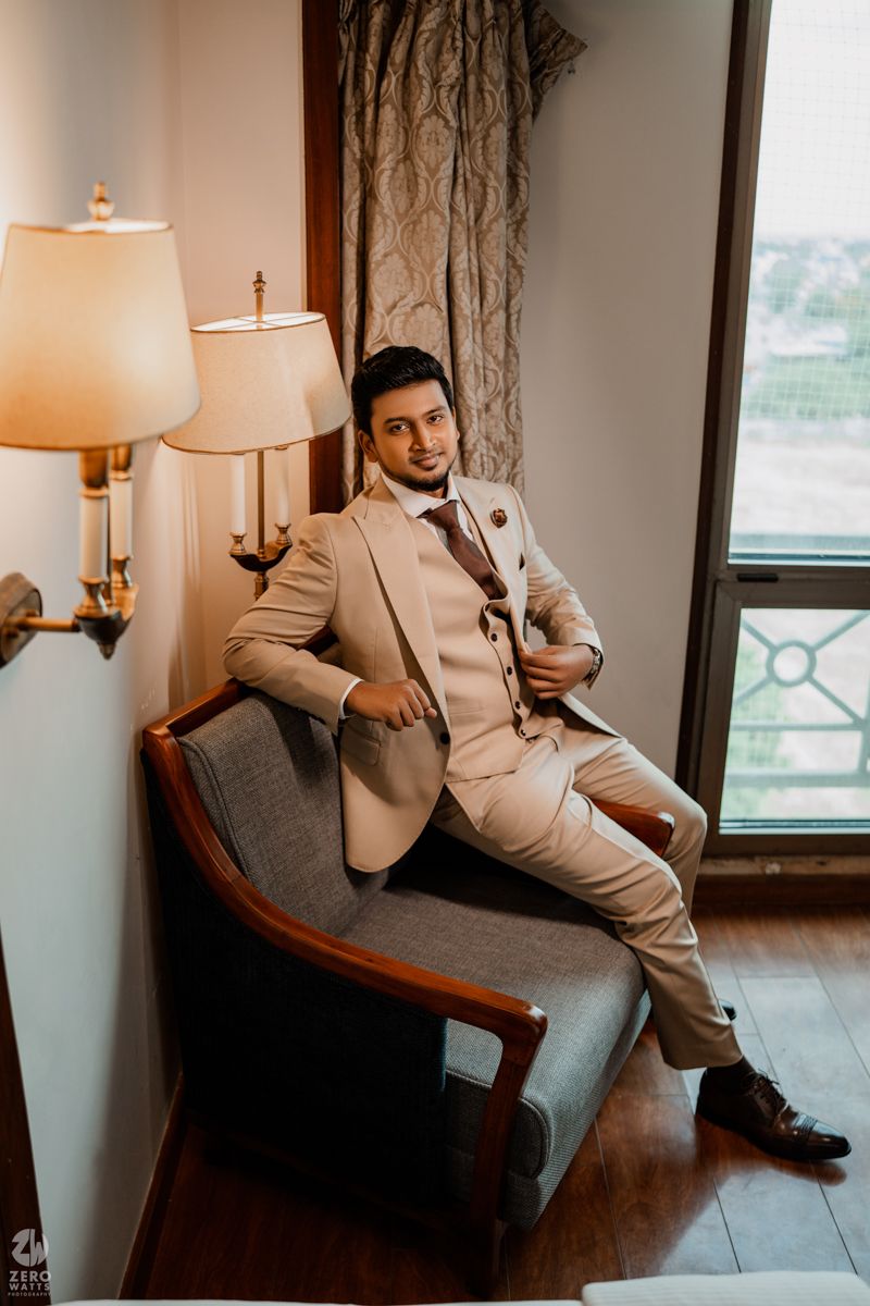 Modern groom lounges in grey chair wearing light beige suit against window with city view.