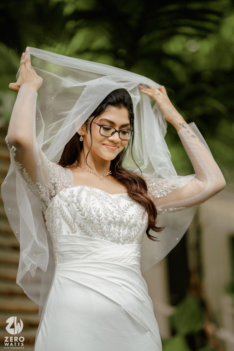 A bride in a white lace wedding dress playfully lifts her veil in a garden setting with natural greenery.