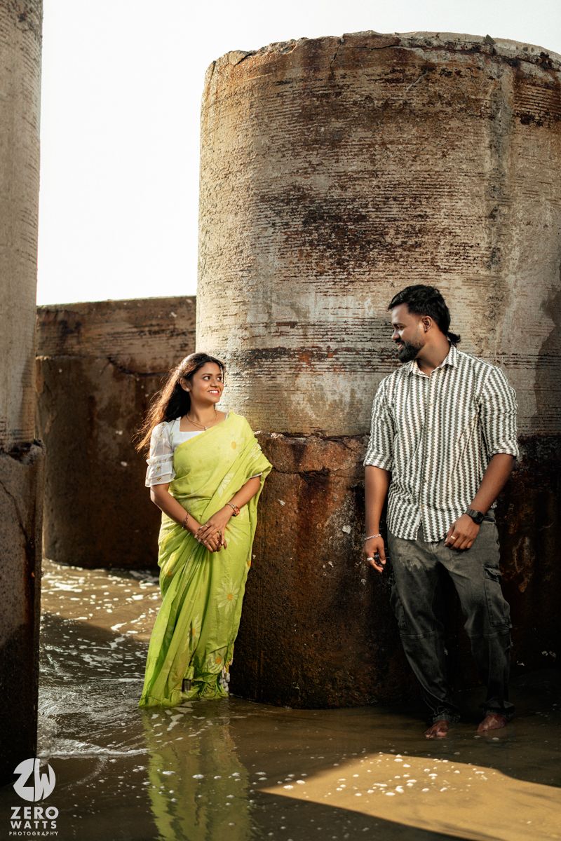 A couple in traditional attire poses romantically against stone pillars near the water's edge.