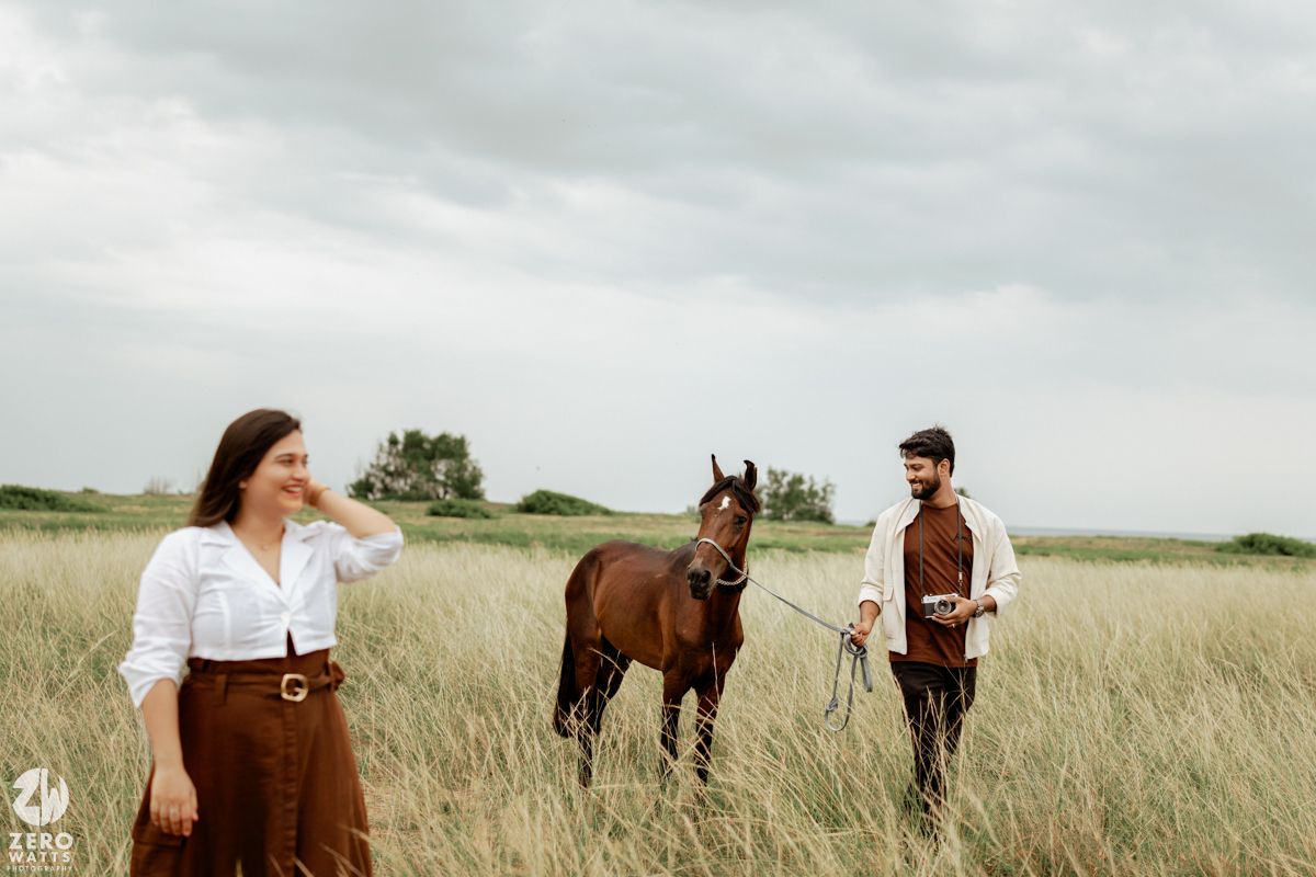 People stand among tall grass in a field with horses visible in the distance under gray skies.