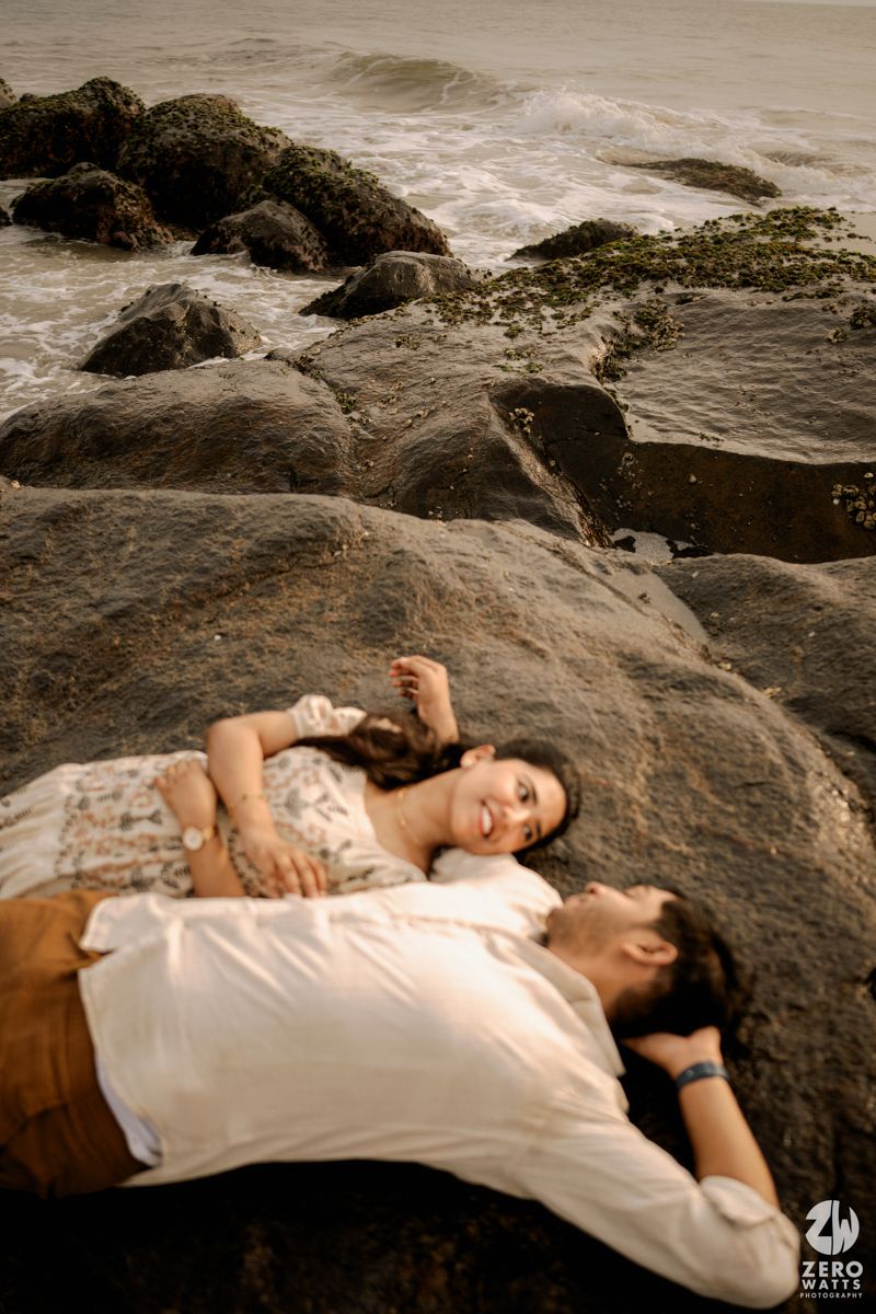 A couple rests together on dark coastal rocks near the ocean during an intimate photoshoot.