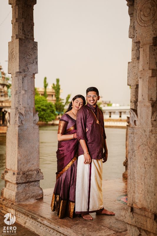 Couple in burgundy attire poses between ornate stone pillars in a temple corridor.