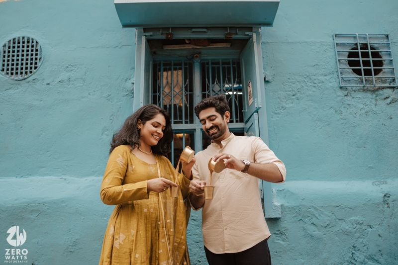 Two people stand together against a turquoise wall wearing casual outfits in warm earth tones.
