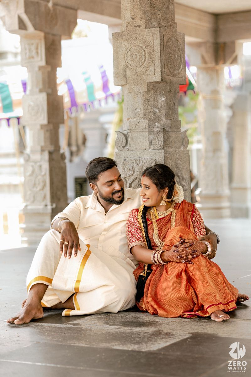 Couple in traditional Indian attire lying on temple floor with colorful decorative flags hanging above them.