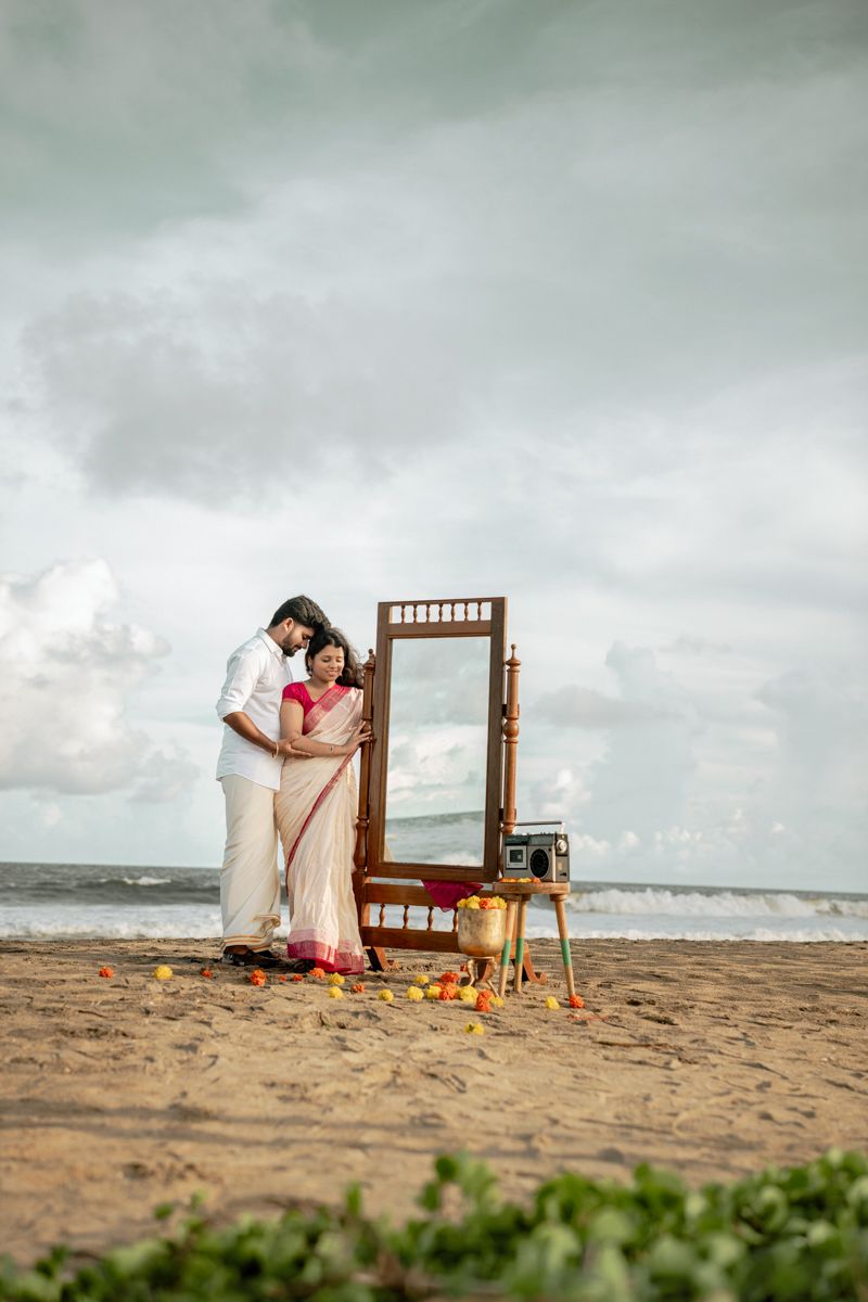 A romantic beach photoshoot setup featuring a wooden mirror frame and vintage decor against cloudy skies.