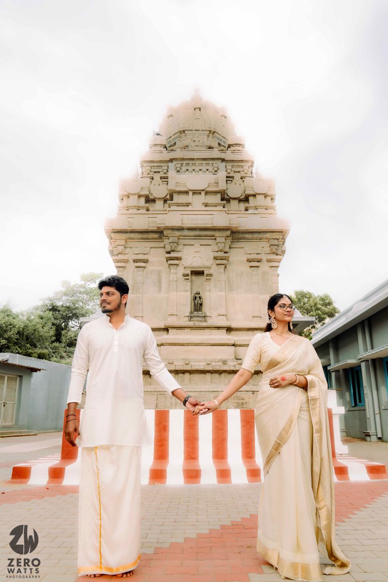 Couple in traditional white attire pose against temple architecture and orange barriers.