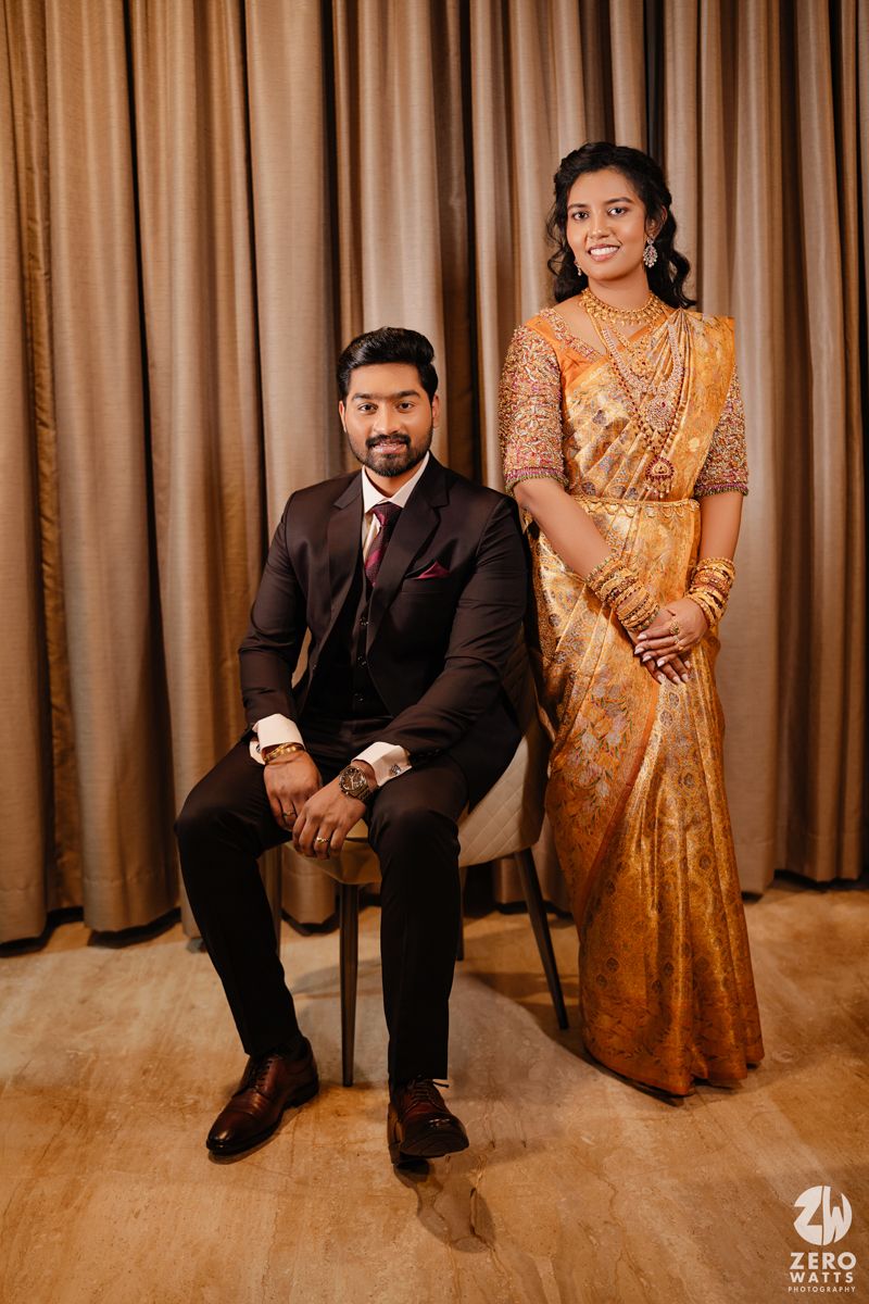 Wedding couple poses together in formal attire with the bride in orange saree and groom in dark suit.