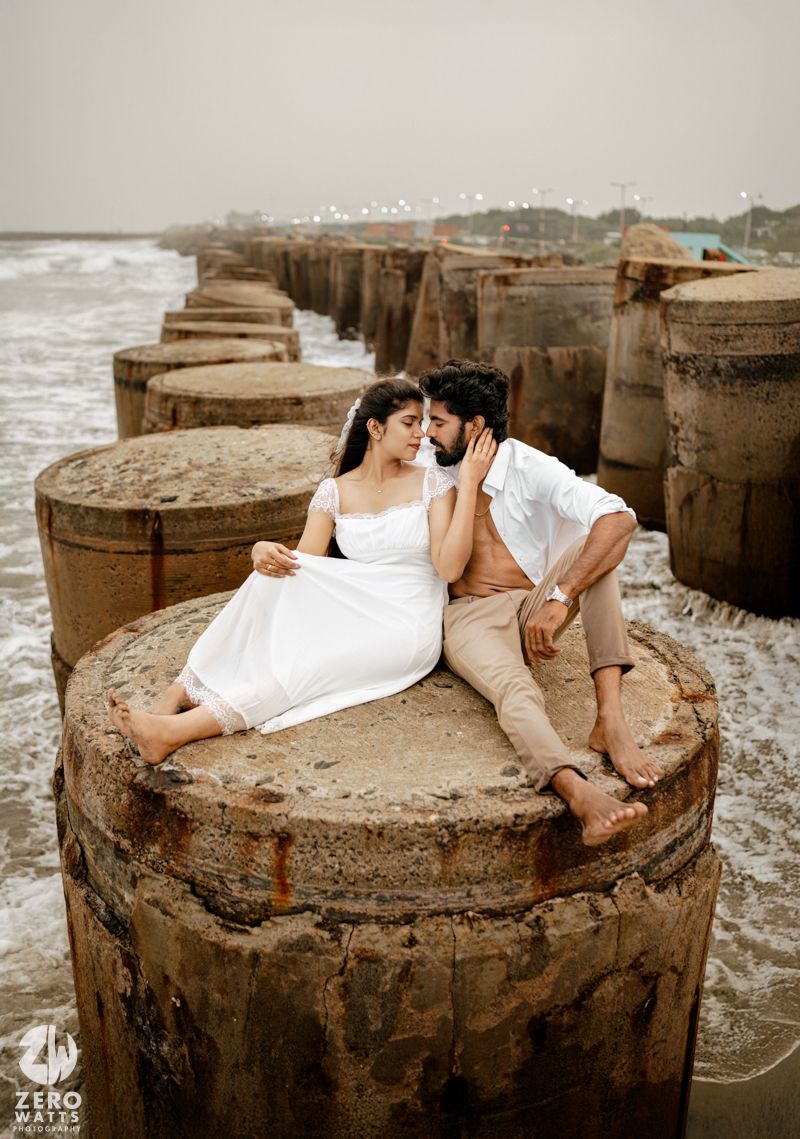 Couple in white and beige sits intimately on concrete pier pillar with ocean waves in background.