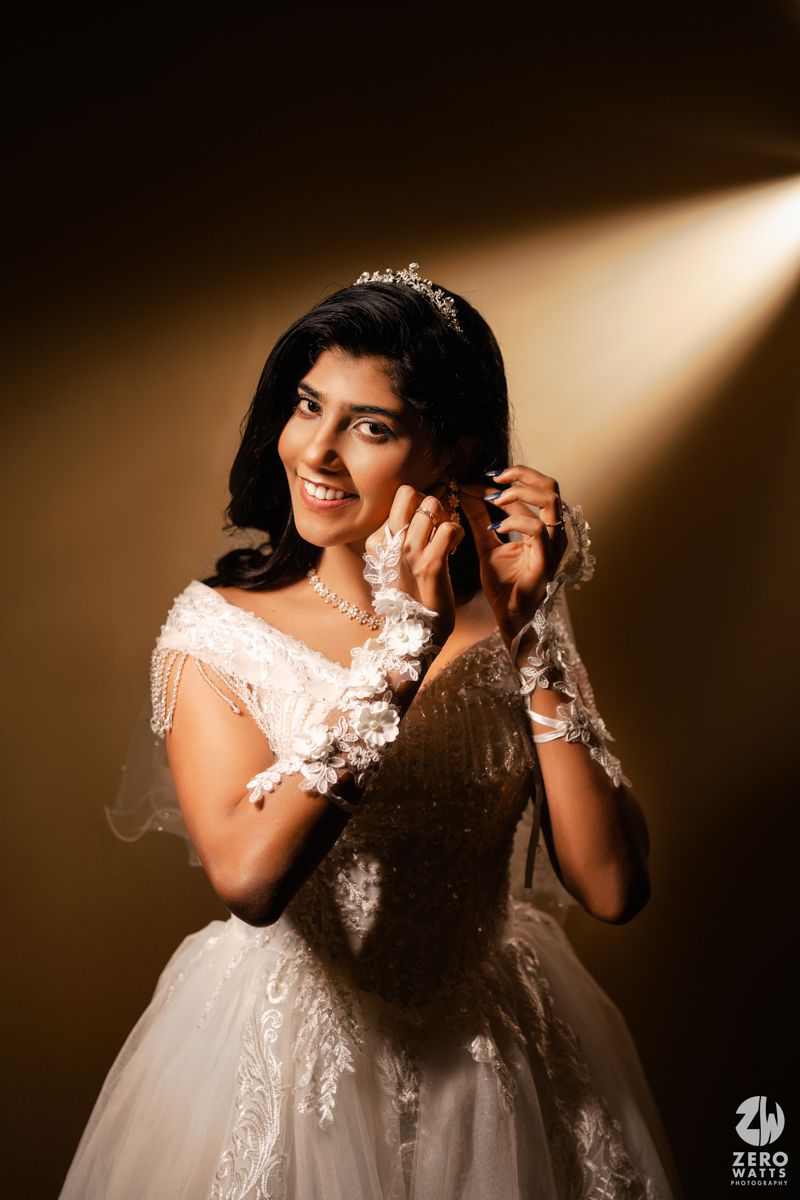 Smiling bride in white ballgown with tiara and lace gloves poses under dramatic spotlight against golden background.