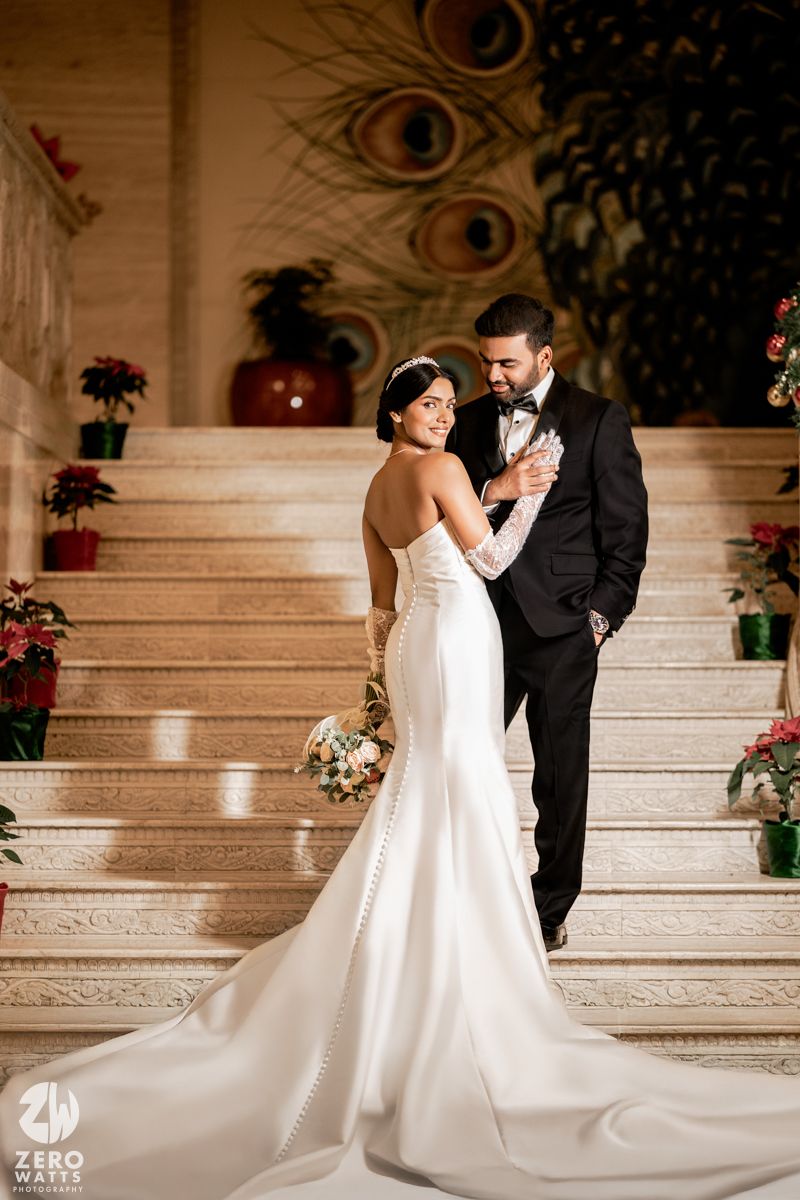 Bride and groom share romantic moment on grand staircase, she in white mermaid gown, he in black tux.