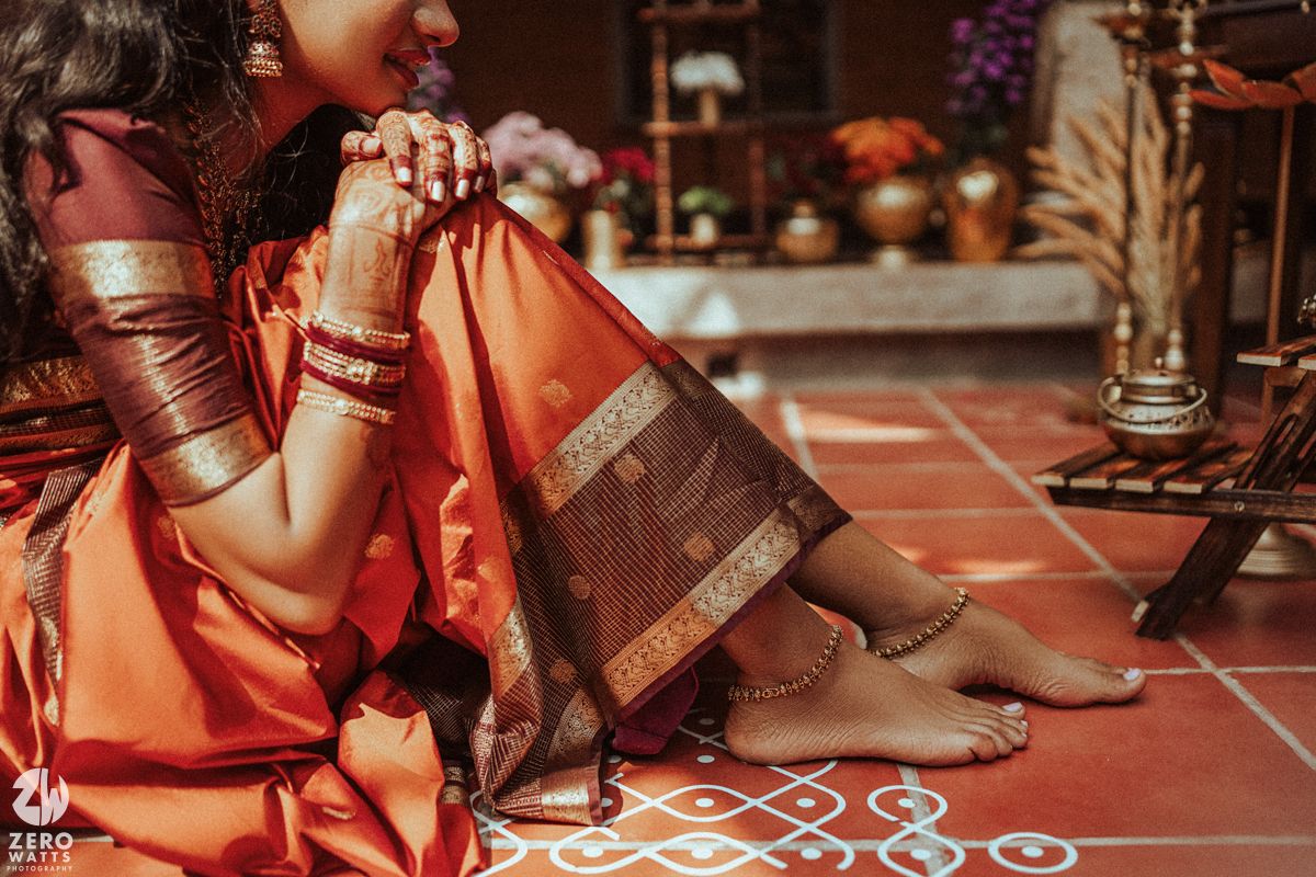 Close-up of bride in orange Kanjivaram silk saree with gold bangles and anklets, seated on kolam-decorated floor.