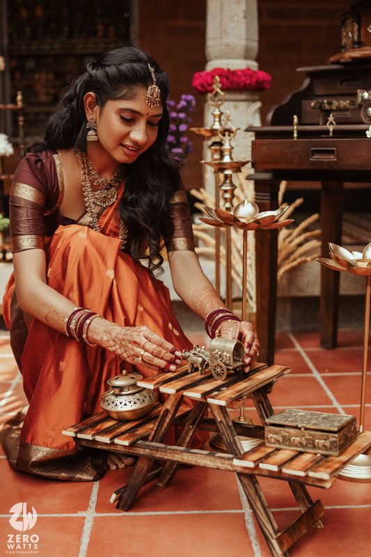 Bride in orange silk saree with mehndi hands arranges items on wooden table during South Indian wedding ritual.
