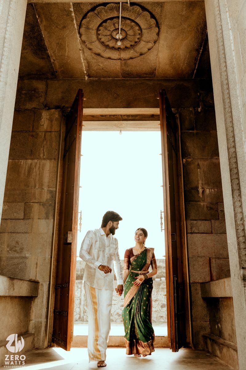 Couple in traditional Indian attire walking through ornate temple doorway, bride in green silk saree, groom in white kurta.