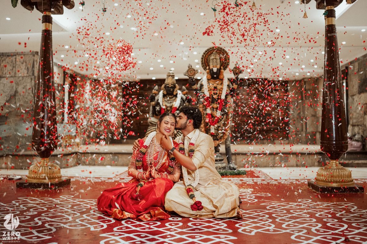 South Indian couple in traditional wedding attire sitting on floor with red and white flower petals falling around them.