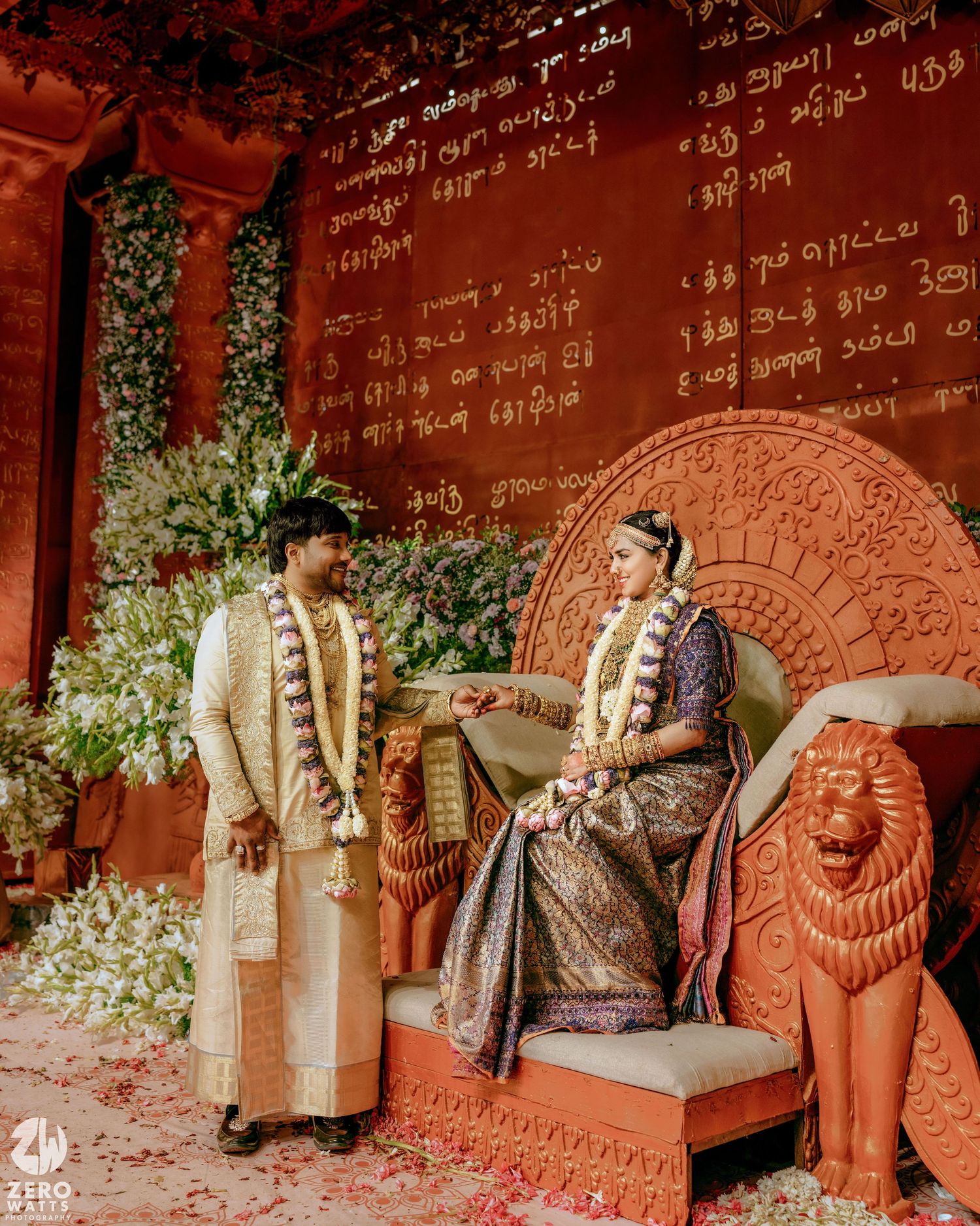 Bride and groom in traditional South Indian wedding attire with flower garlands pose beside a carved lion throne against a decorated wall.
