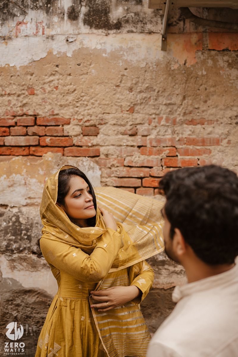A couple in a romantic moment against a weathered brick wall, with one person wearing a mustard yellow dress.