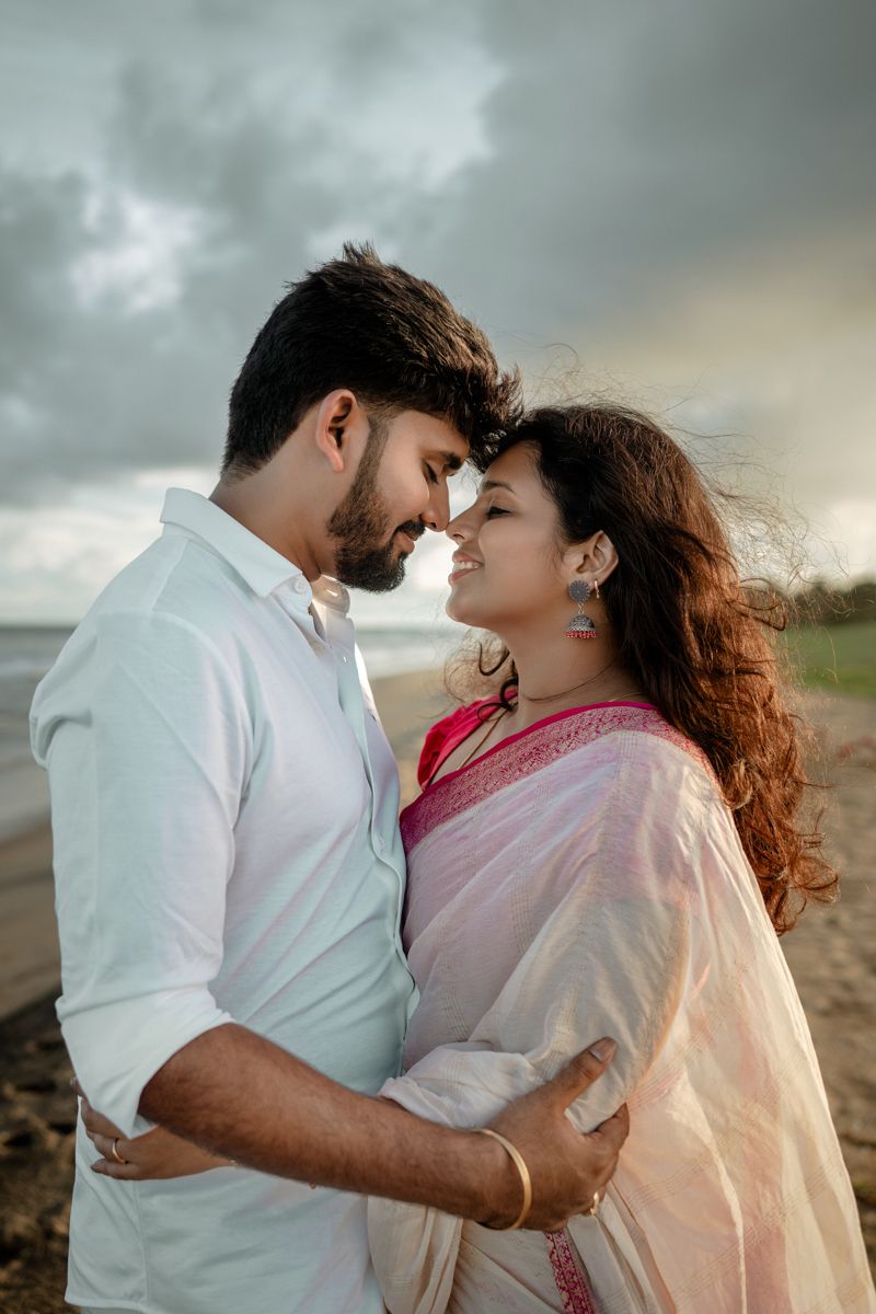 Romantic couple in white and pink outfits share intimate moments on a cloudy beach.