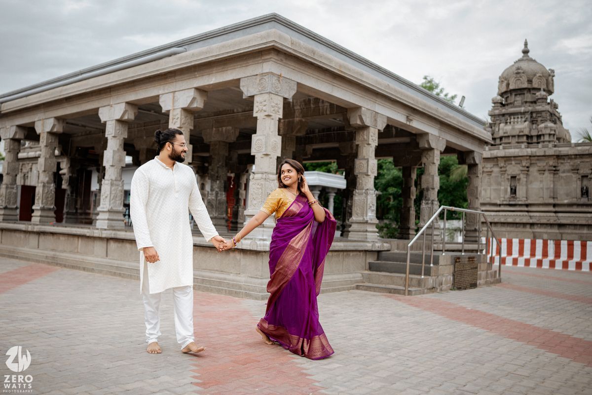 Couple in traditional Indian dress holding hands at temple with stone pillars and dome.