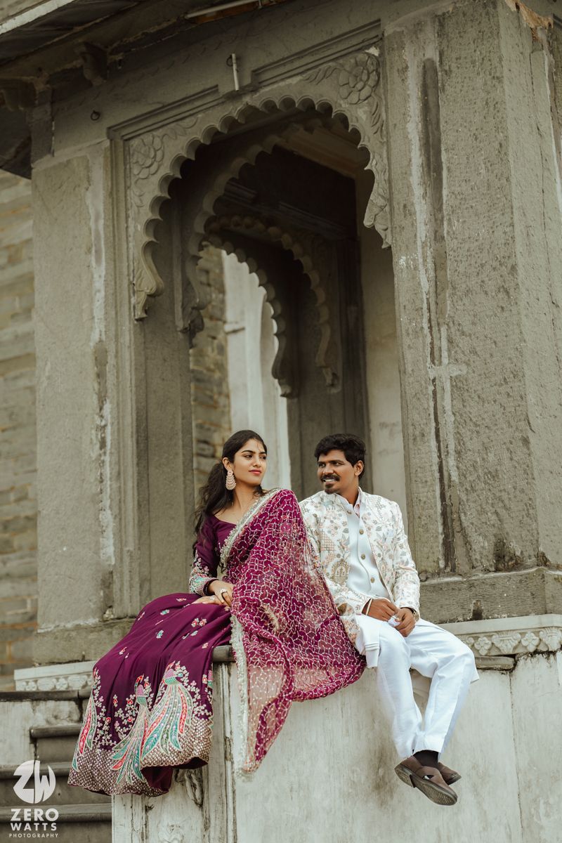 Couple in traditional Indian attire relaxing together on a white stone ledge of a historic building with ornate arches.