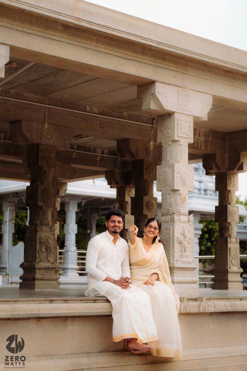 Couple in traditional white attire sits in a temple corridor with ornate stone pillars.